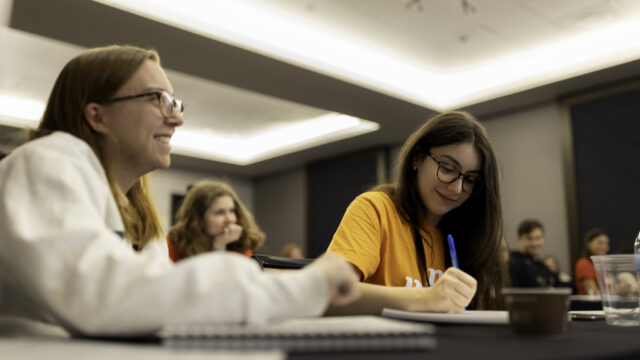Two students are pictured in the foreground with several others blurred in the background. The two students sit at a table with notebooks in front of them; one is smiling at a speaker, who is out of view, and another is looking down at her paper while writing with a blue pen.